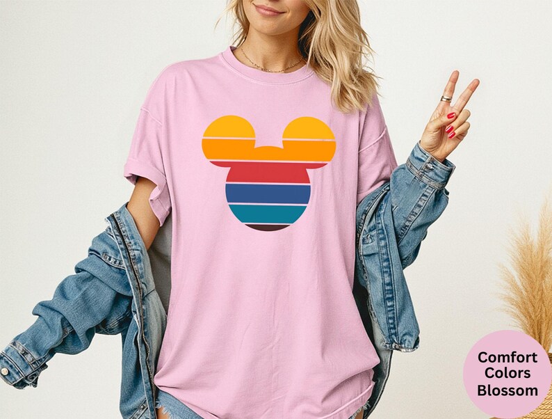 K&ouml;nnte beinhalten: Hellrosa T-Shirt mit einem Retro-Mickey-Mouse-Design. Das Design ist eine Silhouette von Mickeys Kopf mit horizontalen Streifen in Gelb, Orange, Rot, Blau und T&uuml;rkis. Das Shirt ist ein Comfort Colors Blossom.