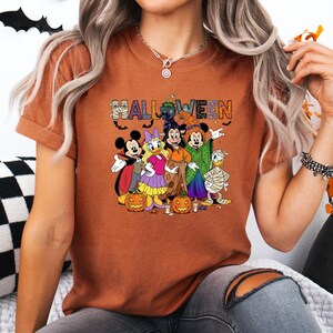 May include: A burnt orange t-shirt featuring a Halloween design with Disney characters. The word "HALLOWEEN" is printed across the top in colorful letters. Mickey Mouse, Daisy Duck, Minnie Mouse, and Donald Duck are dressed in Halloween costumes, with pumpkins in front.