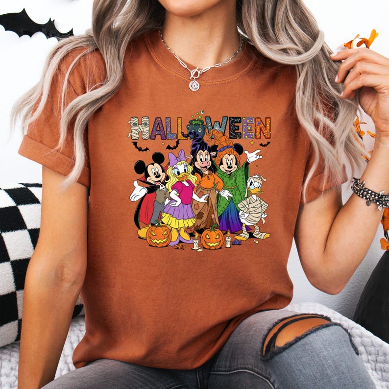 May include: A burnt orange t-shirt featuring a Halloween design with Disney characters. The word "HALLOWEEN" is printed across the top in colorful letters. Mickey Mouse, Daisy Duck, Minnie Mouse, and Donald Duck are dressed in Halloween costumes, with pumpkins in front.