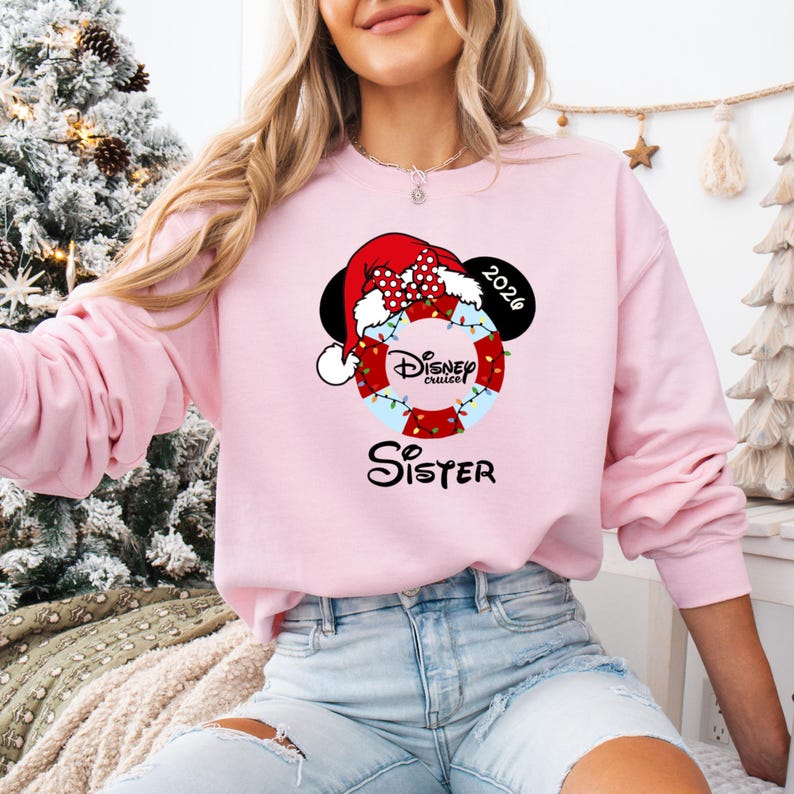 K&ouml;nnte beinhalten: Ein hellrosa Sweatshirt mit einem Disney Cruise-Design. Das Design umfasst Mickey Mouse-Ohren, einen Weihnachtsmannhut und eine Rettungsboje mit den Worten "Disney Cruise" und "Sister". Das Jahr "2026" ist ebenfalls abgebildet.