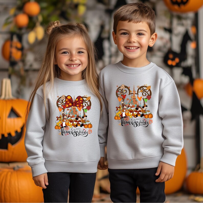 May include: Two children wearing light gray sweatshirts with a Thanksgiving-themed graphic. The graphic features Disney characters, pumpkins, and the text "Happy Thanksgiving". The sweatshirts are long-sleeved.