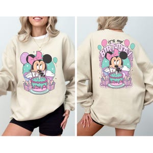 May include: A beige sweatshirt with a graphic print of Minnie Mouse wearing a pink bow and holding a birthday cake. The text "It's my Birthday" is printed on the back of the sweatshirt.