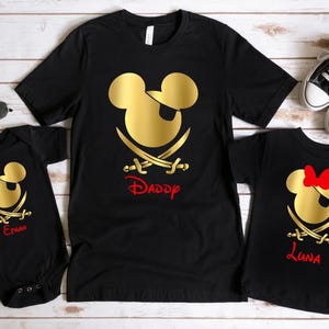 May include: Three black t-shirts with a gold Mickey Mouse pirate design. The shirts have the names "Daddy", "Emma", and "Luna" printed on them.