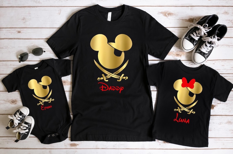 May include: Three black t-shirts with a gold Mickey Mouse pirate design. The shirts have the names "Daddy", "Emma", and "Luna" printed on them.