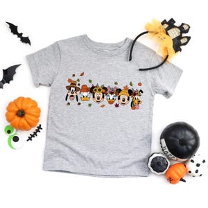 May include: A heather gray t-shirt featuring a graphic of Disney characters in fall attire. The characters include Goofy, Donald Duck, Mickey Mouse, Daisy Duck, Minnie Mouse, and Pluto. The shirt is decorated with fall leaves and pumpkins.