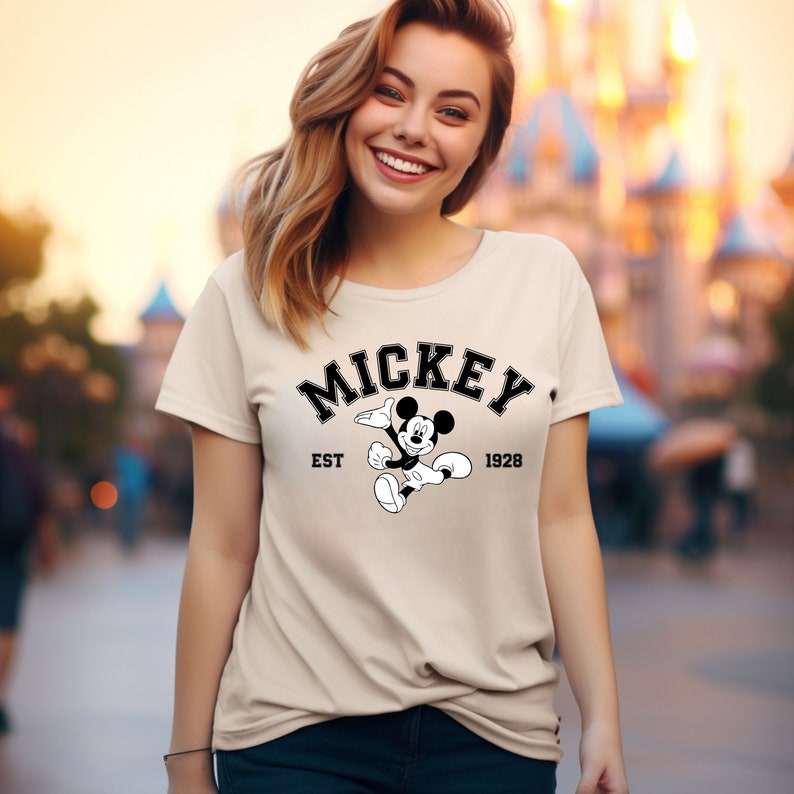 May include: A beige t-shirt with a black and white graphic of Mickey Mouse. The graphic includes the text "Mickey" in a black arch above Mickey Mouse, "EST" to the left of Mickey Mouse, and "1928" to the right of Mickey Mouse.