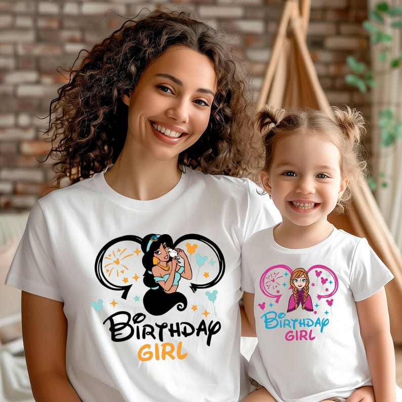 May include: Two white t-shirts with a Disney princess theme. The larger shirt features Jasmine from Aladdin with the text "Birthday Girl". The smaller shirt features Elsa from Frozen with the text "Birthday Girl".