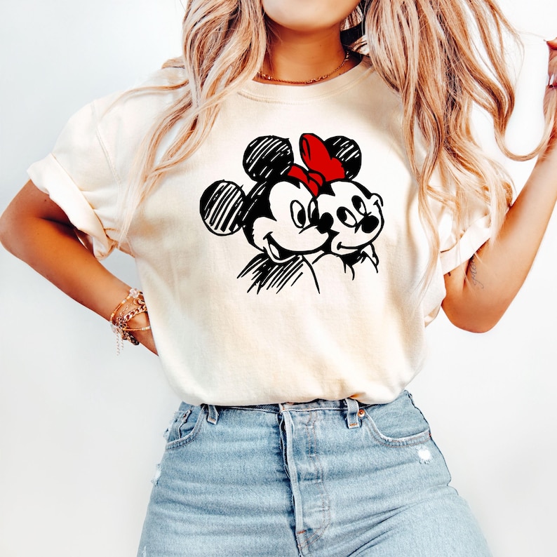 May include: Cream-colored t-shirt featuring a black and white sketch of Mickey and Minnie Mouse. Minnie wears a red bow. The design is centered on the shirt, which has rolled-up sleeves and is paired with blue jeans.