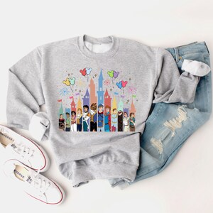 Puede incluir: Una sudadera gris con una ilustraci&oacute;n colorida de pr&iacute;ncipes de Disney frente a un castillo con globos y fuegos artificiales. La sudadera est&aacute; colocada sobre una superficie blanca con un par de zapatillas blancas y unos vaqueros azules.