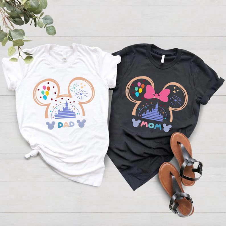 May include: Two white t-shirts with a black t-shirt in the background. The white t-shirts have a Mickey Mouse head design with the words "DAD" and "MOM" printed on them. The design includes a castle, balloons, fireworks, and stars.