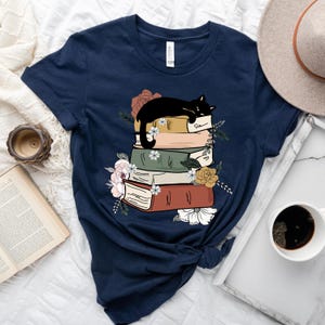May include: A navy blue t-shirt with a black cat sleeping on a stack of books. The books are decorated with flowers and have a vintage aesthetic.