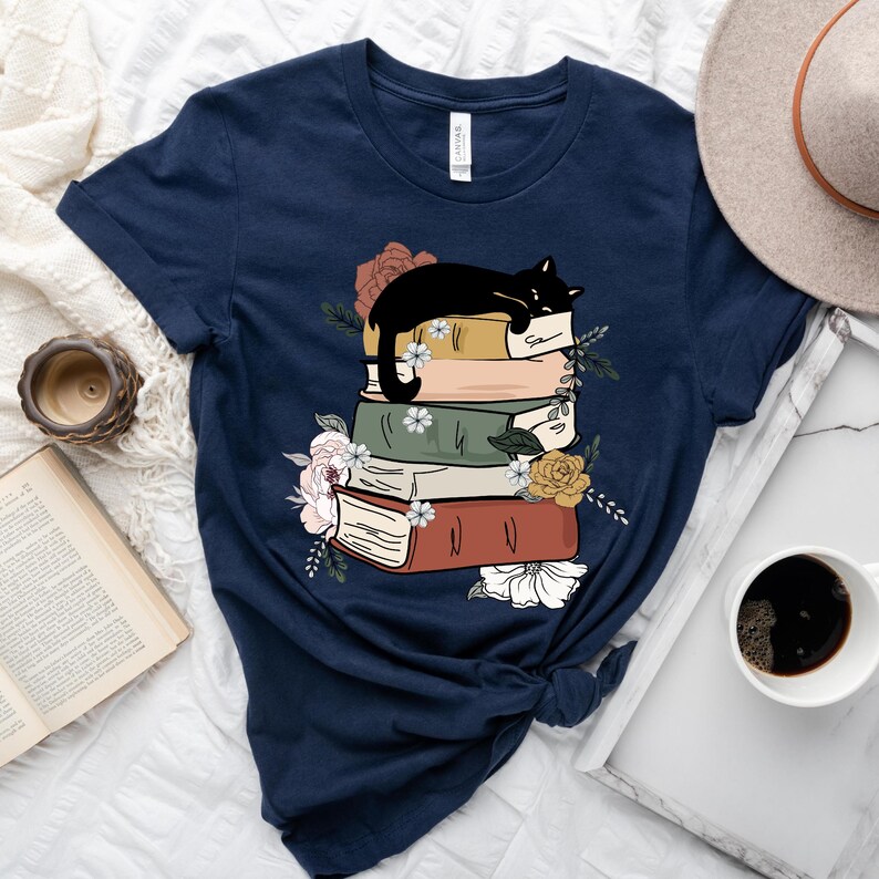 May include: A navy blue t-shirt with a black cat sleeping on a stack of books. The books are decorated with flowers and have a vintage aesthetic.