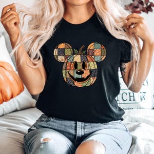May include: Black t-shirt featuring a pumpkin design with Mickey Mouse ears. The pumpkin is made of patchwork squares in various colors, with a smiling face. The shirt is a casual style, suitable for Halloween or fall.