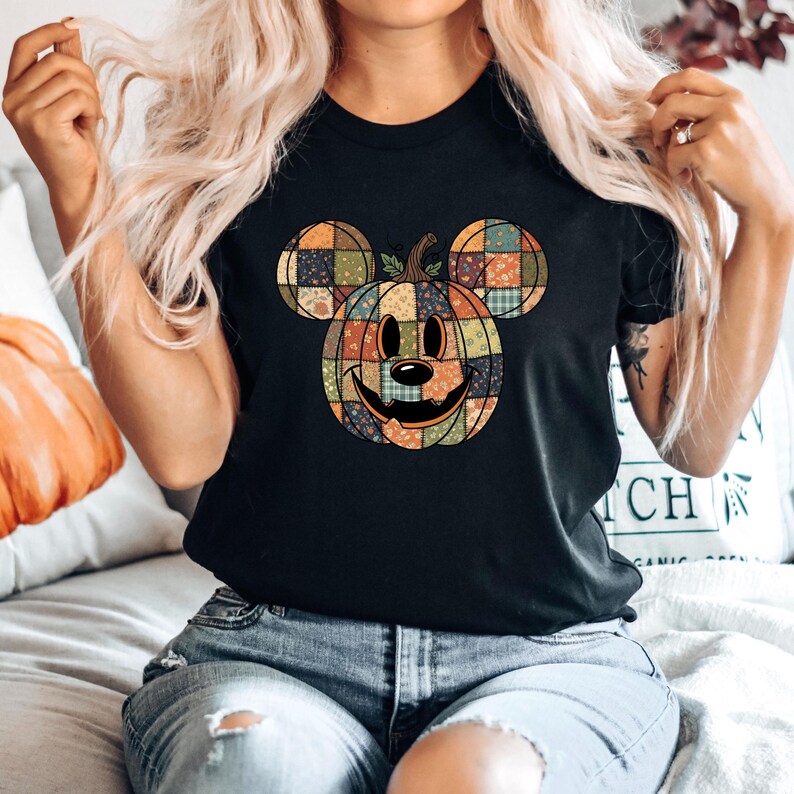May include: Black t-shirt featuring a pumpkin design with Mickey Mouse ears. The pumpkin is made of patchwork squares in various colors, with a smiling face. The shirt is a casual style, suitable for Halloween or fall.