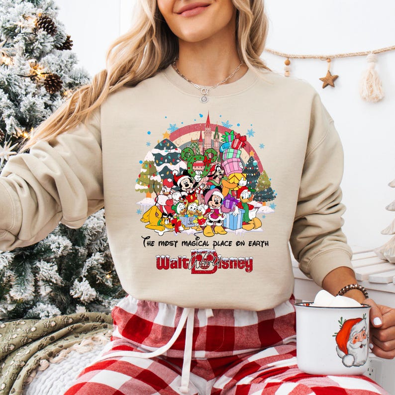 Op de afbeelding: Beige sweatshirt met een kleurrijke afbeelding van Disney-personages, een kasteel en een regenboog. De tekst "The most magical place on earth" en "Walt Disney" staan eronder. De persoon draagt een rood-witte geruite broek.