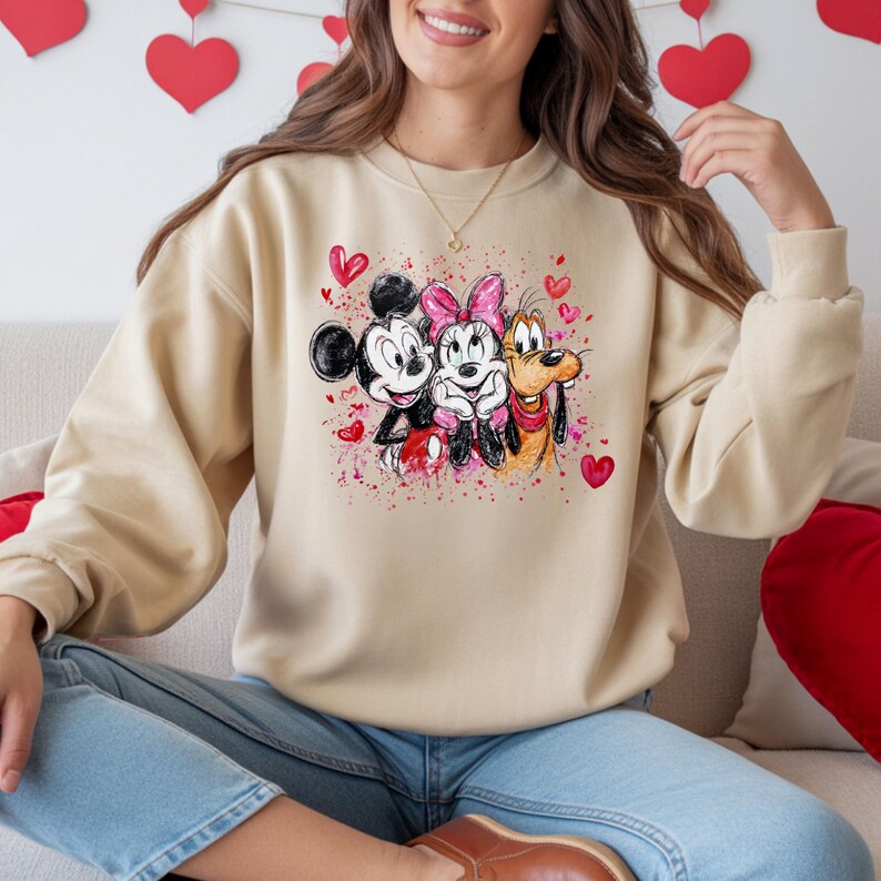 May include: Beige sweatshirt featuring a colorful graphic of Mickey Mouse, Minnie Mouse, and Pluto. The design includes pink hearts and watercolor-style splashes. The sweatshirt has long sleeves and a crew neck.