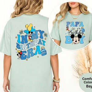 Op de afbeelding: Lichtgroene T-shirts met Mickey Mouse-ontwerpen. E&eacute;n shirt zegt "In My Birthday Era" met Mickey Mouse en een taart. De andere zegt "Papa of the Birthday Boy" met Mickey Mouse. Beide shirts hebben blauwe en gele accenten.