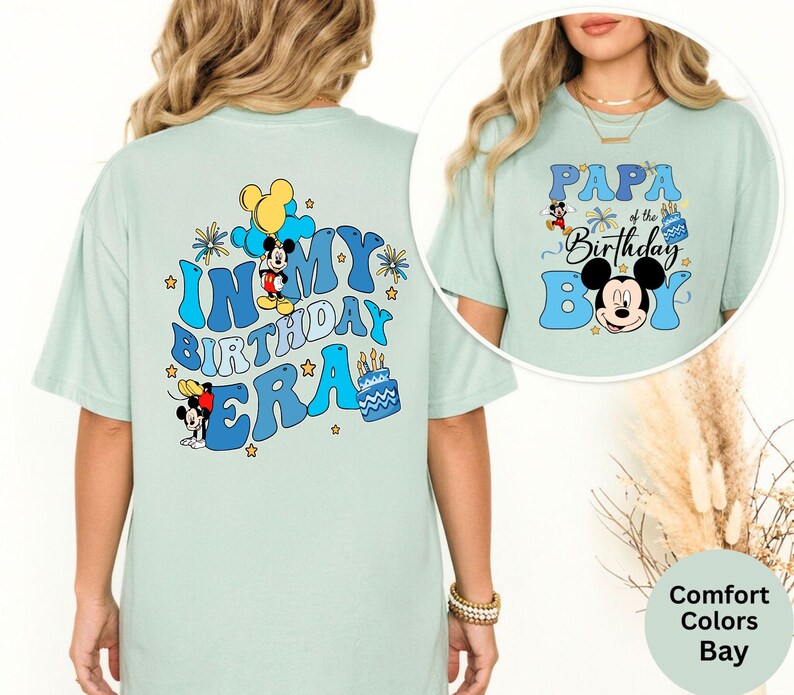 Op de afbeelding: Lichtgroene T-shirts met Mickey Mouse-ontwerpen. E&eacute;n shirt zegt "In My Birthday Era" met Mickey Mouse en een taart. De andere zegt "Papa of the Birthday Boy" met Mickey Mouse. Beide shirts hebben blauwe en gele accenten.