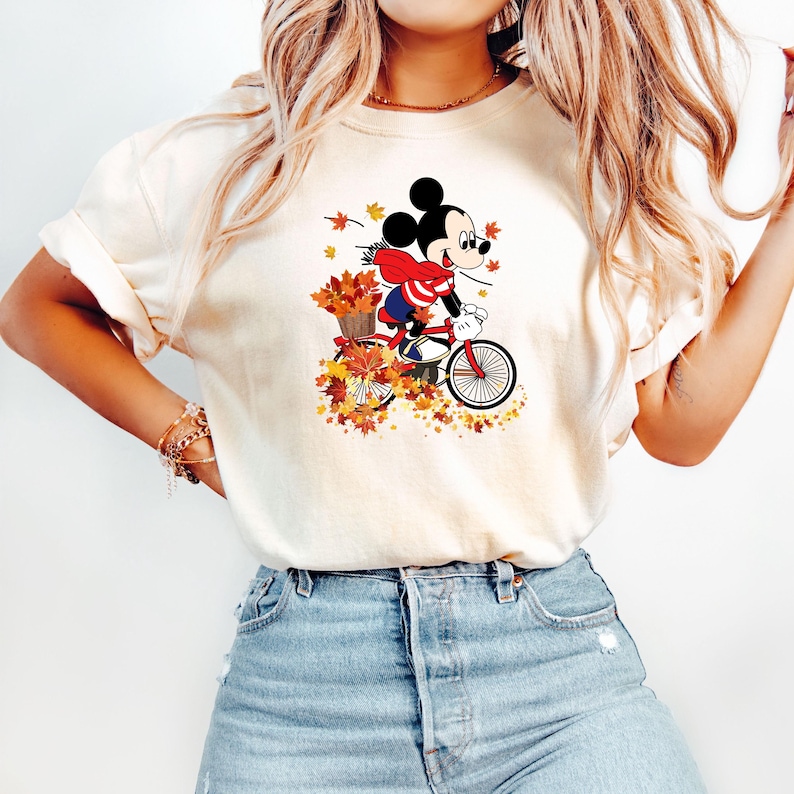 May include: Cream-colored t-shirt featuring a graphic of Mickey Mouse riding a red bicycle through autumn leaves. Mickey wears a red scarf and is carrying a basket of fall foliage. The design is set against a backdrop of falling leaves.