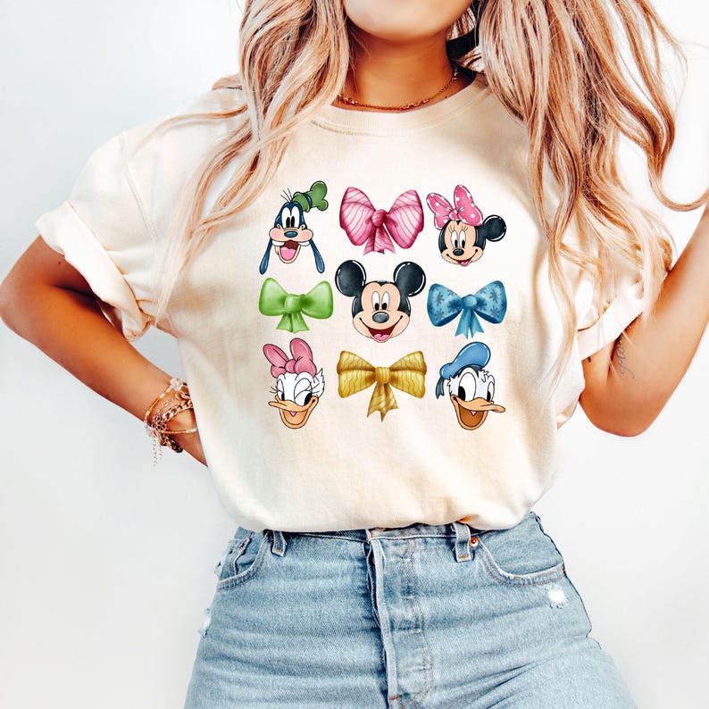 May include: A white t-shirt with a colorful graphic print of Disney characters, including Mickey Mouse, Minnie Mouse, Donald Duck, Goofy, and Daisy Duck. Each character is wearing a bow.