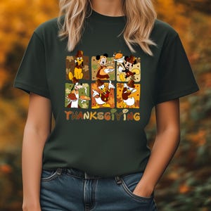 May include: Dark green t-shirt featuring a Thanksgiving-themed graphic. The graphic includes cartoon characters like Mickey Mouse, Donald Duck, and Goofy, each in a festive setting. The word "THANKSGIVING" is displayed in colorful letters.