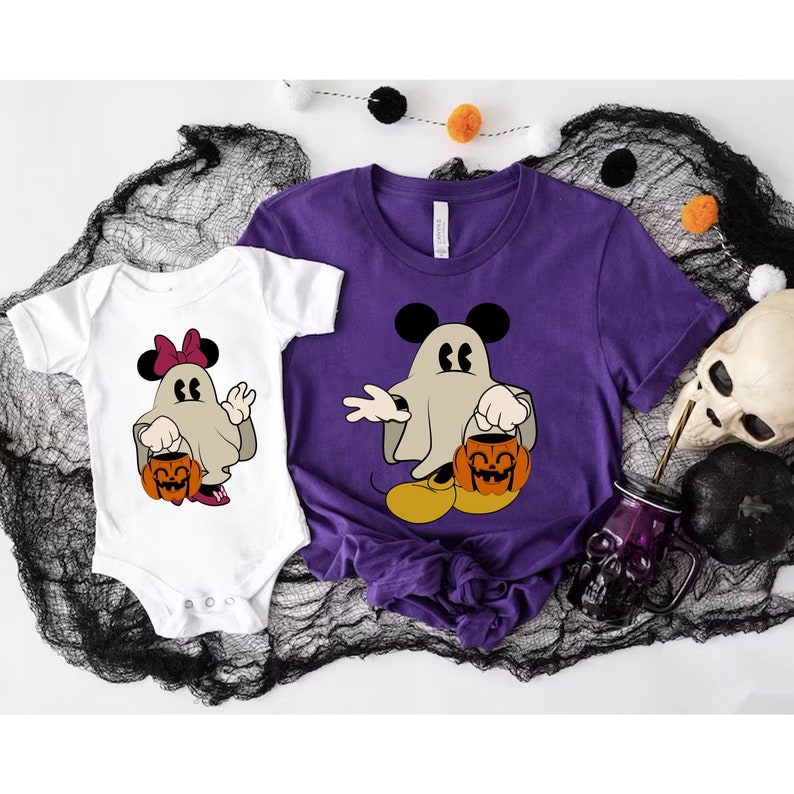 Pode incluir: Uma camiseta roxa e um body branco com um personagem de fantasma de desenho animado com orelhas do Mickey Mouse, segurando um balde de ab&oacute;bora. O fantasma est&aacute; usando um len&ccedil;ol. O body tem um la&ccedil;o rosa. Roupas com tema de Halloween.