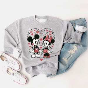 May include: A gray sweatshirt with a graphic print of Mickey Mouse and Minnie Mouse holding hands. The graphic is surrounded by red hearts and a heart-shaped outline.