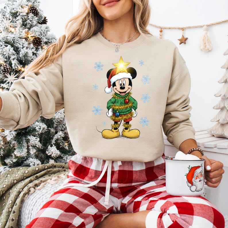 Pode incluir: Camisola bege com gola redonda com um gr&aacute;fico do Mickey Mouse com tema natalino. Mickey est&aacute; vestido com um su&eacute;ter verde, cal&ccedil;&otilde;es vermelhos e um gorro de Papai Noel, rodeado por flocos de neve. O gr&aacute;fico inclui uma estrela dourada e sapatos amarelos.