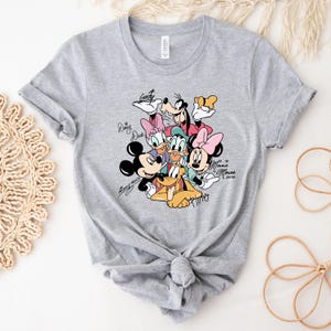 May include: A light gray t-shirt featuring a colorful graphic of Disney characters including Mickey Mouse, Minnie Mouse, Donald Duck, and Goofy. The characters' signatures are printed around the image.