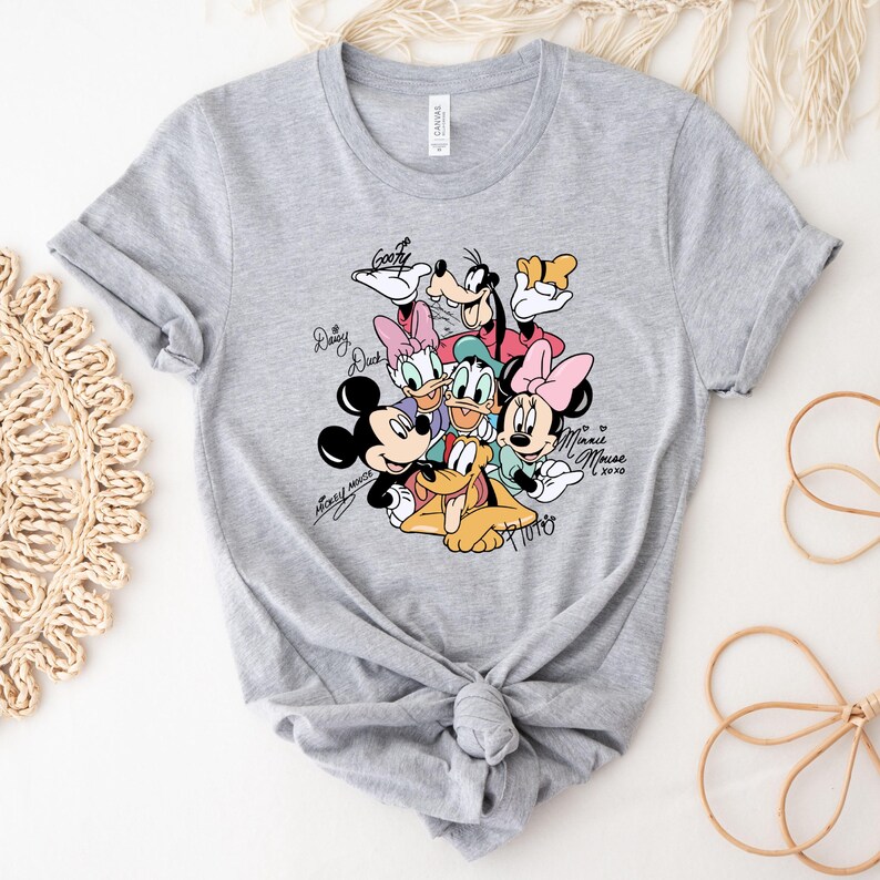 May include: A light gray t-shirt featuring a colorful graphic of Disney characters including Mickey Mouse, Minnie Mouse, Donald Duck, and Goofy. The characters' signatures are printed around the image.