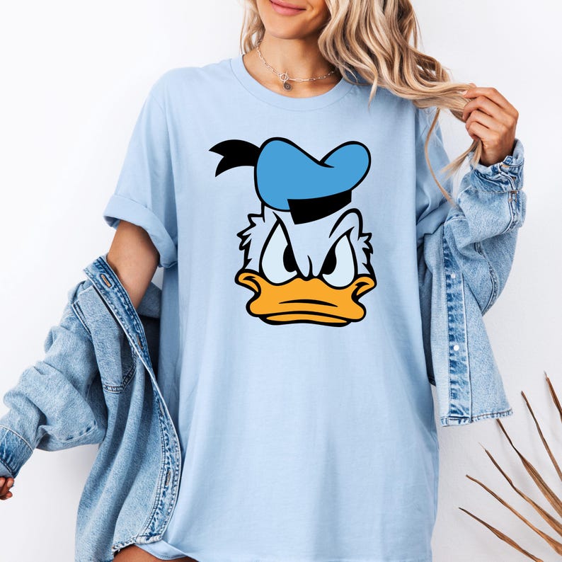 May include: Light blue t-shirt featuring a cartoon graphic of Donald Duck's face. The design includes a blue sailor hat, a yellow beak, and an angry expression. The shirt has short sleeves and a relaxed fit.