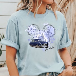 Puede incluir: Una camiseta azul claro con un coche azul y un boceto de orejas de Mickey Mouse con un castillo en su interior. El texto "Doc Hudson" est&aacute; impreso debajo del coche.