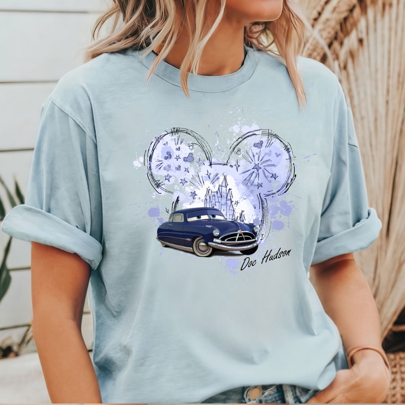 Puede incluir: Una camiseta azul claro con un coche azul y un boceto de orejas de Mickey Mouse con un castillo en su interior. El texto "Doc Hudson" est&aacute; impreso debajo del coche.