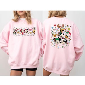 May include: A light pink sweatshirt featuring a festive design with Disney characters. The front displays Mickey, Minnie, Donald, Daisy, Goofy, and Pluto in Santa hats, strung with Christmas lights. The back shows a similar design.