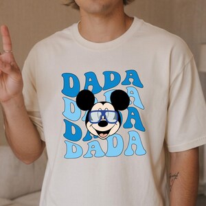 K&ouml;nnte beinhalten: Cremefarbenes T-Shirt mit Retro-Design. Das Design zeigt das Wort "DADA" in Hellblau, dreifach gestapelt, und eine Cartoon-Mickey-Mouse mit Brille. Das T-Shirt hat eine lockere Passform.