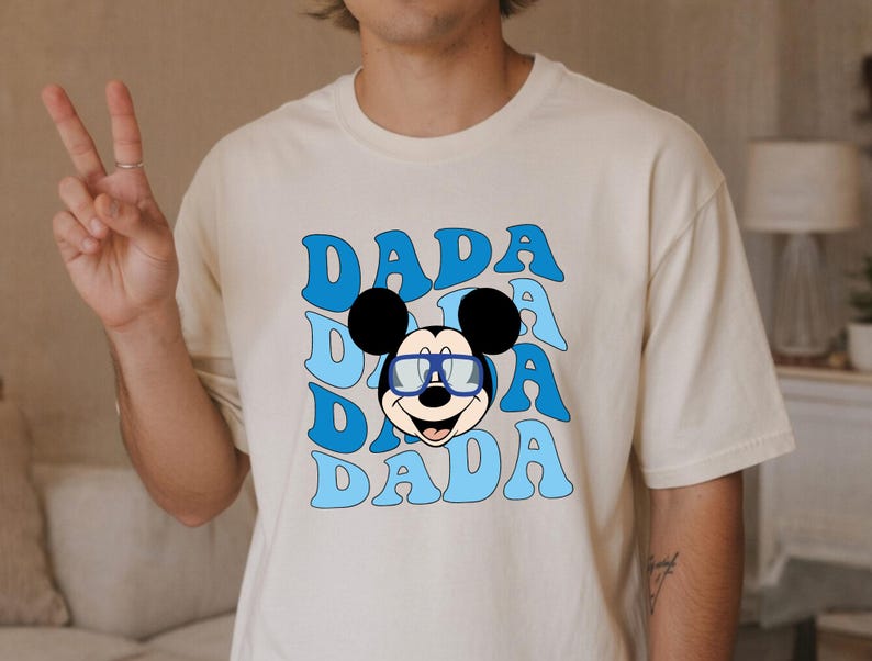 K&ouml;nnte beinhalten: Cremefarbenes T-Shirt mit Retro-Design. Das Design zeigt das Wort "DADA" in Hellblau, dreifach gestapelt, und eine Cartoon-Mickey-Mouse mit Brille. Das T-Shirt hat eine lockere Passform.
