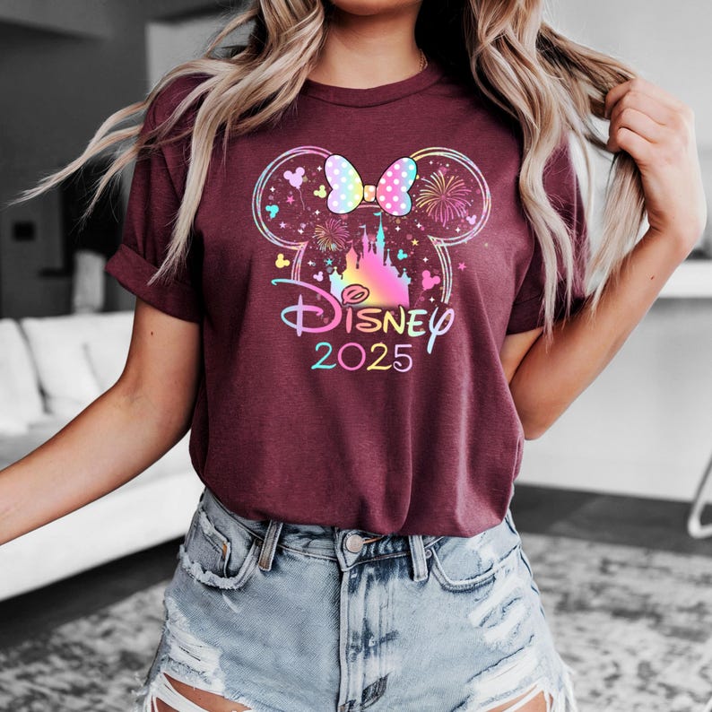 May include: A maroon t-shirt with a colorful graphic of Mickey Mouse ears, a bow, fireworks, and Cinderella's castle. The text "DISNEY 2025" is printed below the graphic.
