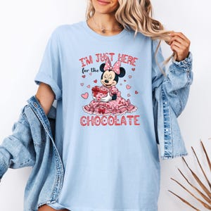 May include: Light blue t-shirt featuring a graphic of Minnie Mouse holding a heart-shaped gift box. The text reads "I'M JUST HERE for the CHOCOLATE" in red. The design includes pink hearts and a pink bow.