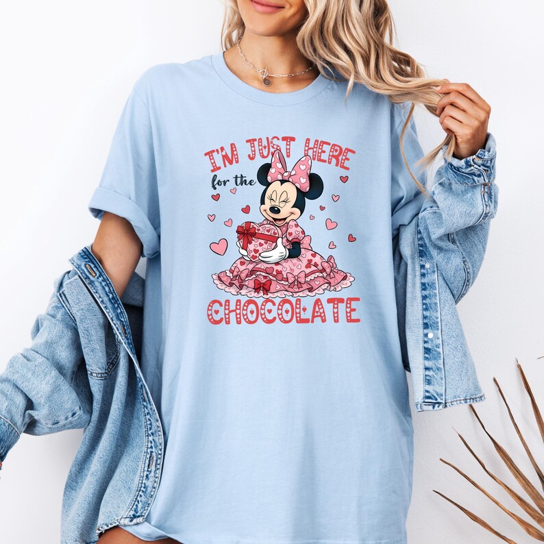 May include: Light blue t-shirt featuring a graphic of Minnie Mouse holding a heart-shaped gift box. The text reads "I'M JUST HERE for the CHOCOLATE" in red. The design includes pink hearts and a pink bow.