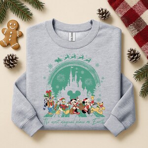 以下が含まれることがあります： 緑色の城、雪の結晶、クリスマス衣装のディズニーキャラクターのグラフィックが特徴のライトグレーのスウェットシャツ。「The most magical place on Earth」の文字が下部に印刷されています。お祝いの休日の装飾がスウェットシャツを囲んでいます。