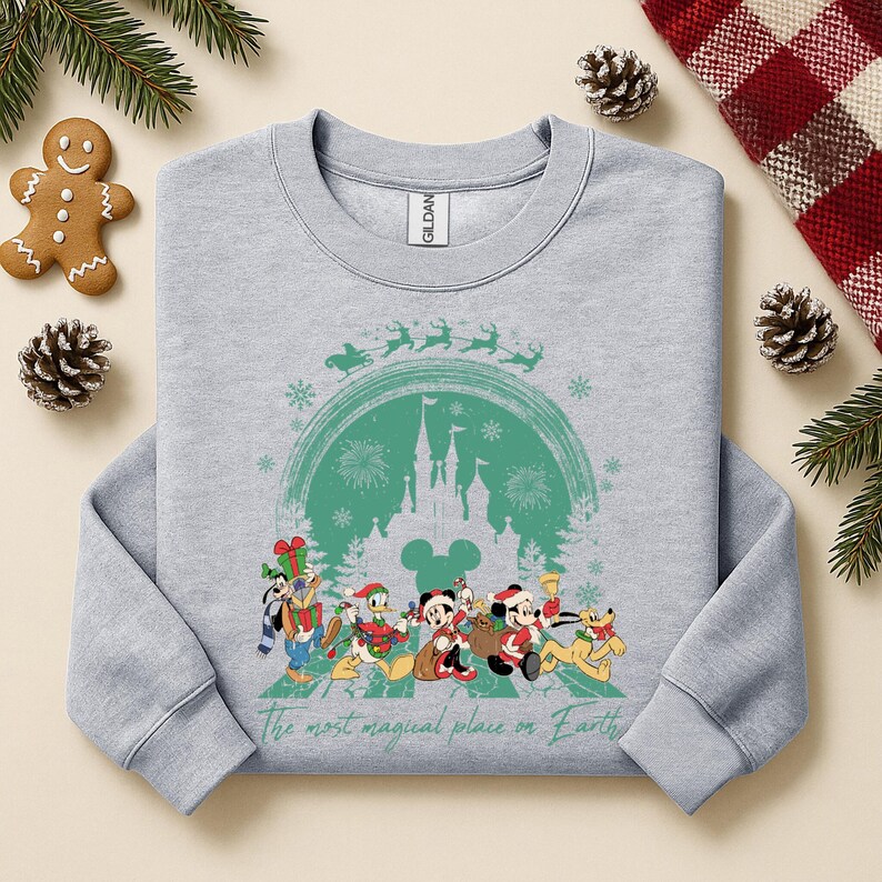 以下が含まれることがあります： 緑色の城、雪の結晶、クリスマス衣装のディズニーキャラクターのグラフィックが特徴のライトグレーのスウェットシャツ。「The most magical place on Earth」の文字が下部に印刷されています。お祝いの休日の装飾がスウェットシャツを囲んでいます。