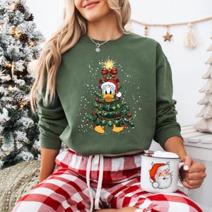 May include: Green sweatshirt featuring a Christmas tree design with Donald Duck wearing a Santa hat. The tree is decorated with red ornaments and lights. The person is wearing red and white plaid pajama pants and holding a mug with a Santa Claus graphic.