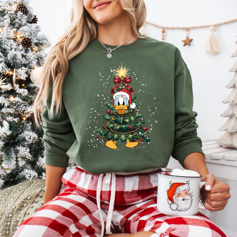 May include: Green sweatshirt featuring a Christmas tree design with Donald Duck wearing a Santa hat. The tree is decorated with red ornaments and lights. The person is wearing red and white plaid pajama pants and holding a mug with a Santa Claus graphic.