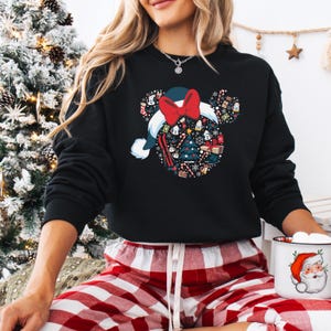May include: Black sweatshirt with a festive design of a Santa hat with a red bow and Mickey Mouse ears. The hat is filled with Christmas-themed icons. The person is wearing red and white plaid pajama bottoms and holding a Santa mug.