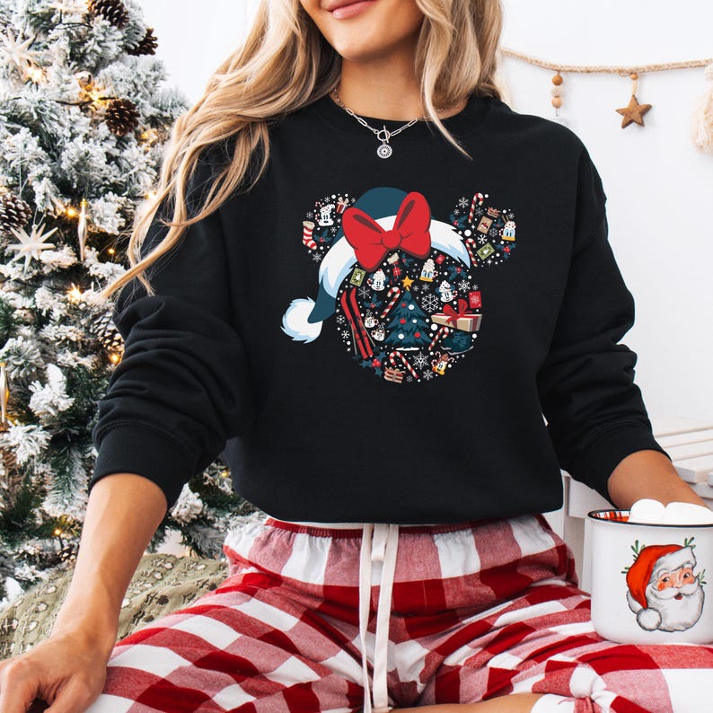 May include: Black sweatshirt with a festive design of a Santa hat with a red bow and Mickey Mouse ears. The hat is filled with Christmas-themed icons. The person is wearing red and white plaid pajama bottoms and holding a Santa mug.