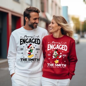 Puede incluir: Una pareja lleva su&eacute;teres navide&ntilde;os a juego con un dise&ntilde;o de Mickey Mouse y Minnie Mouse. Los su&eacute;teres son blancos y rojos con el texto "Merry & Engaged" y "EST. 2024" en ellos. Los su&eacute;teres tambi&eacute;n tienen el texto "The Smith" y "First Christmas Mr. and Mrs."