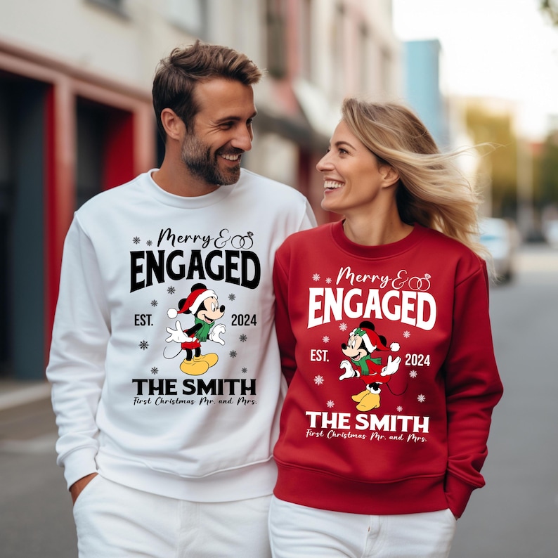 Puede incluir: Una pareja lleva su&eacute;teres navide&ntilde;os a juego con un dise&ntilde;o de Mickey Mouse y Minnie Mouse. Los su&eacute;teres son blancos y rojos con el texto "Merry & Engaged" y "EST. 2024" en ellos. Los su&eacute;teres tambi&eacute;n tienen el texto "The Smith" y "First Christmas Mr. and Mrs."