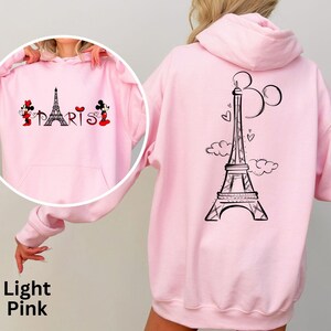 K&ouml;nnte beinhalten: Hellrosa Kapuzenpullover mit Fronttasche und Kapuze. Auf der Vorderseite befindet sich eine schwarze Eiffelturm-Grafik mit dem Wort "PARIS" und Mickey und Minnie Mouse. Auf der R&uuml;ckseite befindet sich ein gr&ouml;&szlig;eres Eiffelturm-Design mit Mickey Mouse Ohren.