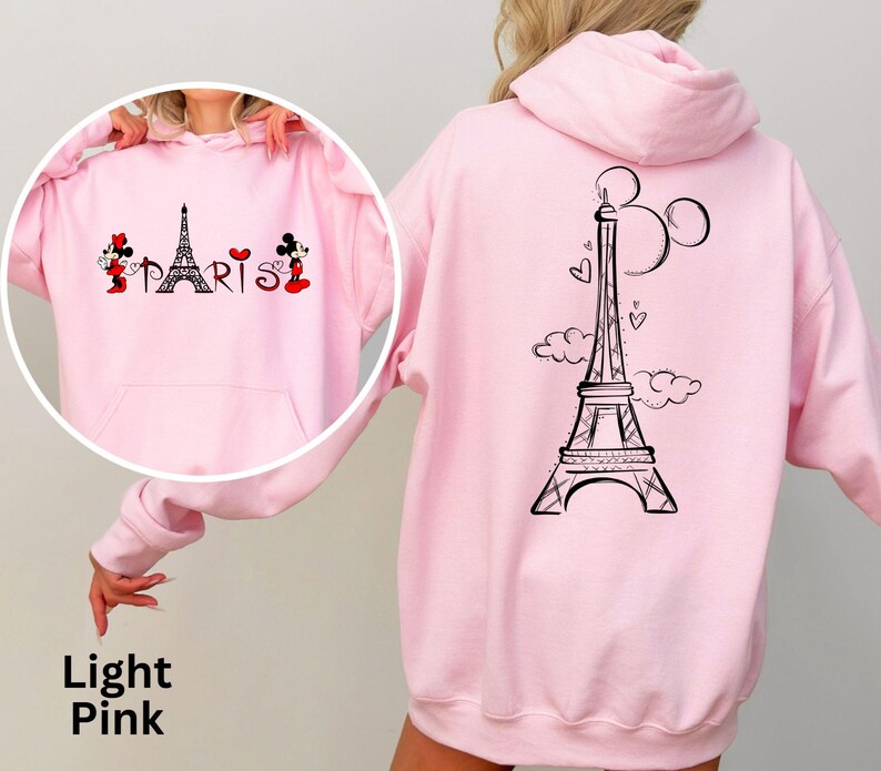 K&ouml;nnte beinhalten: Hellrosa Kapuzenpullover mit Fronttasche und Kapuze. Auf der Vorderseite befindet sich eine schwarze Eiffelturm-Grafik mit dem Wort "PARIS" und Mickey und Minnie Mouse. Auf der R&uuml;ckseite befindet sich ein gr&ouml;&szlig;eres Eiffelturm-Design mit Mickey Mouse Ohren.