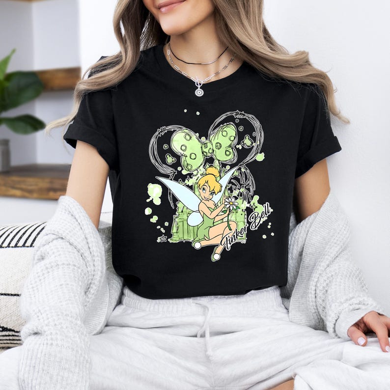 May include: Black t-shirt with a green Tinker Bell graphic. The graphic features Tinker Bell sitting on a branch with a flower in her hand. The graphic is surrounded by green and white paint splatters.